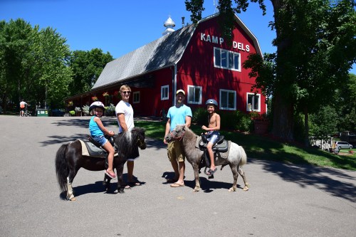A family stands with two ponies in front of a red barn on a sunny day. Two children are riding the ponies.