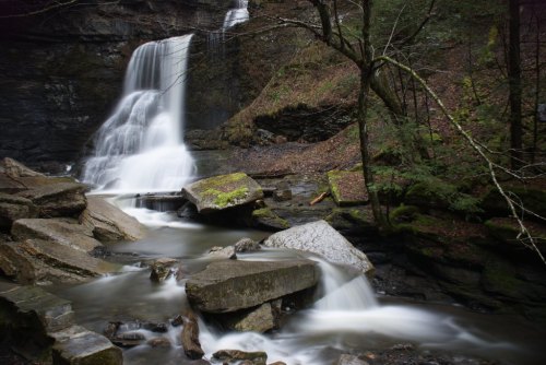 A serene waterfall cascades over rocks, surrounded by lush greenery and a tranquil stream.