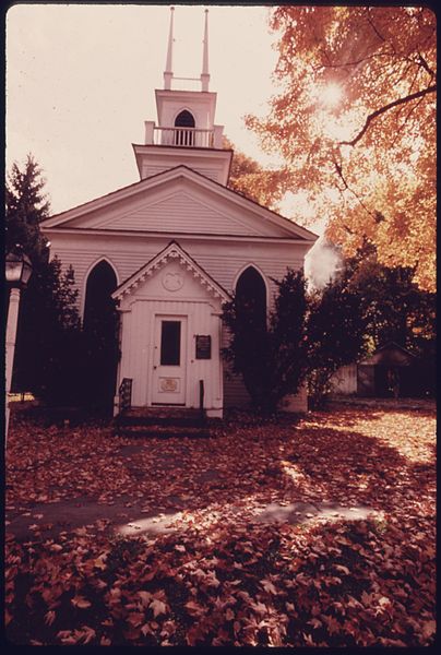 A quaint white church surrounded by colorful autumn leaves and trees, with a clear sky in the background.