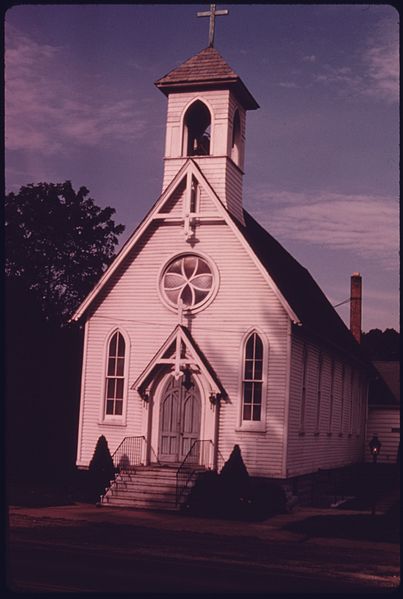 A white wooden church with a bell tower, circular window, and steps leading to the entrance, set against a cloudy sky.