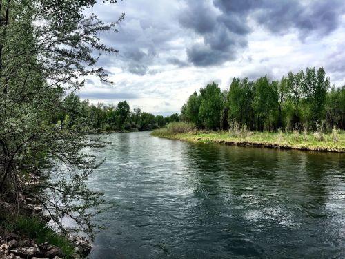 A serene river flows through lush greenery under a cloudy sky, surrounded by trees and natural landscape.