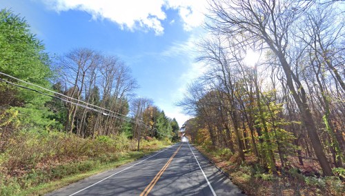 A scenic road lined with trees, showcasing autumn colors under a bright blue sky with scattered clouds.