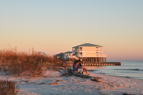 A serene beach scene at sunset, featuring a couple sitting on a log near a colorful house on stilts by the water.