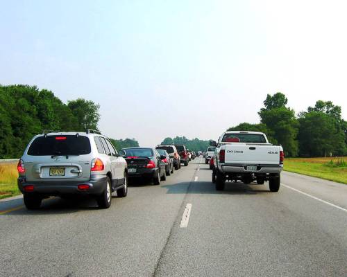 Traffic jam on a highway with multiple vehicles lined up, surrounded by green trees and open fields.