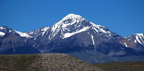 A snow-capped mountain rises against a clear blue sky, surrounded by rugged terrain and lower hills.