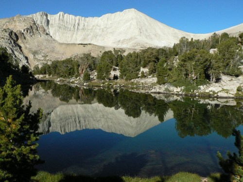 A serene mountain landscape with a clear lake reflecting the peaks and trees under a blue sky.