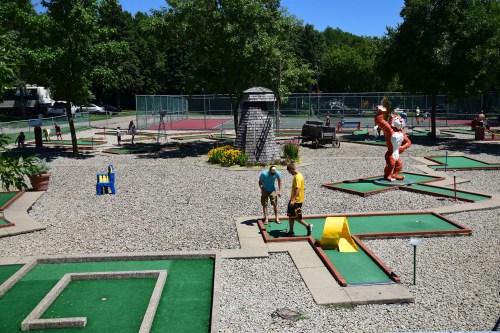 A mini-golf course with players, colorful obstacles, and trees in the background on a sunny day.