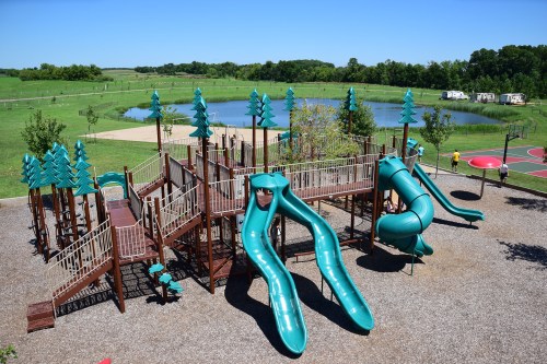 A large playground with green slides, surrounded by trees, near a pond and sports courts on a sunny day.