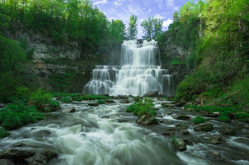 A cascading waterfall surrounded by lush green trees and rocky terrain, with a river flowing in the foreground.
