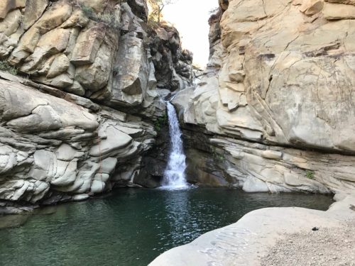 A serene waterfall cascades into a clear pool, surrounded by rugged rock formations and natural beauty.