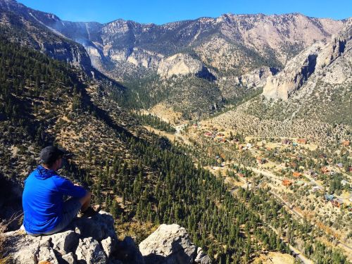 A person sits on a rocky ledge overlooking a valley filled with trees and small houses, surrounded by mountains.