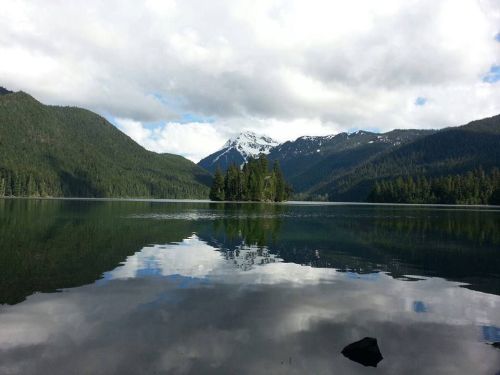 A serene lake surrounded by mountains, with a small island and reflections of clouds and trees on the water's surface.