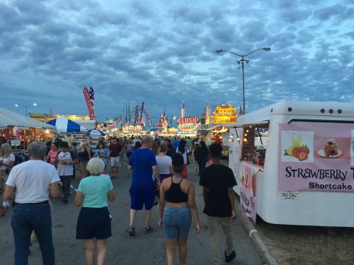 A crowd walks through a fairground with colorful food stalls and a cloudy sky in the background.
