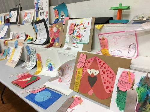 A display of colorful children's artwork on a table, featuring various drawings and crafts with tags.
