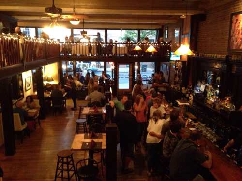 A lively bar scene with patrons socializing, a two-level layout, and warm lighting creating a cozy atmosphere.