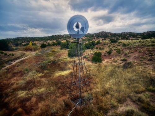 A windmill stands in a grassy landscape under a cloudy sky, surrounded by rolling hills and sparse vegetation.