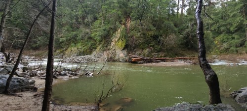 A serene river flows through a forested area, surrounded by rocky banks and lush greenery.