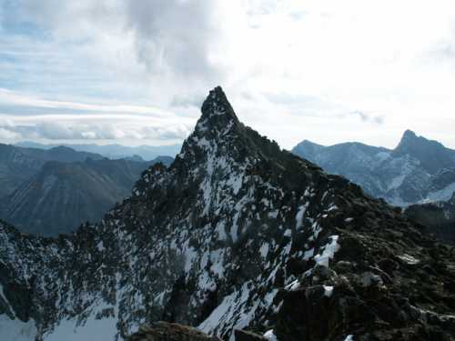 A jagged mountain peak rises above a snowy landscape, surrounded by rugged terrain and distant mountains under a cloudy sky.