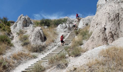 A steep wooden staircase leads up a rocky hillside, with hikers ascending under a clear blue sky.
