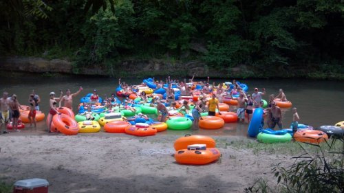 A lively group of people enjoying a river day, surrounded by colorful inflatable tubes on a sandy shore.