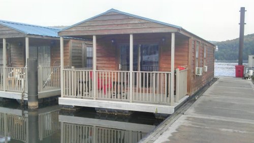 A wooden houseboat with a blue roof and porch, docked beside another houseboat on calm water.