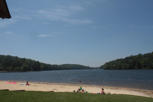 A serene lakeside scene with a sandy beach, people relaxing, and lush green hills in the background under a clear blue sky.