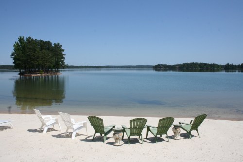 A serene lakeside view with white and green chairs on sandy shore, surrounded by calm water and trees in the distance.