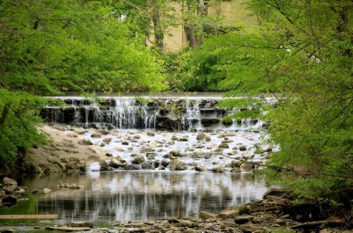 A serene waterfall cascades over rocks, surrounded by lush green trees and a calm river reflecting the scenery.