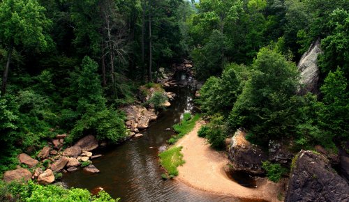 A serene river winding through lush green trees and rocky banks, with a sandy area along the water's edge.