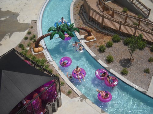 Aerial view of a lazy river with people floating on pink inner tubes, surrounded by palm trees and greenery.