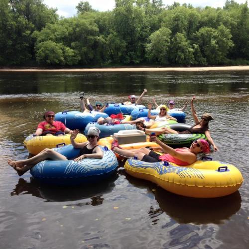 A group of people relaxing on colorful inner tubes in a river, surrounded by lush greenery and a sunny sky.