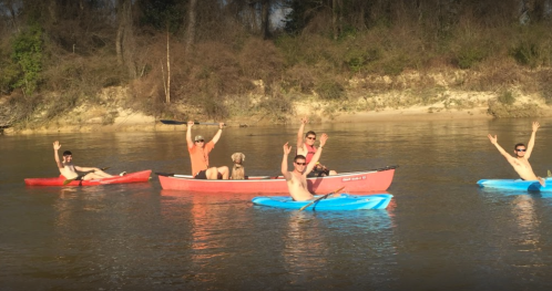 A group of six people in kayaks and a canoe, smiling and waving on a calm river surrounded by trees.