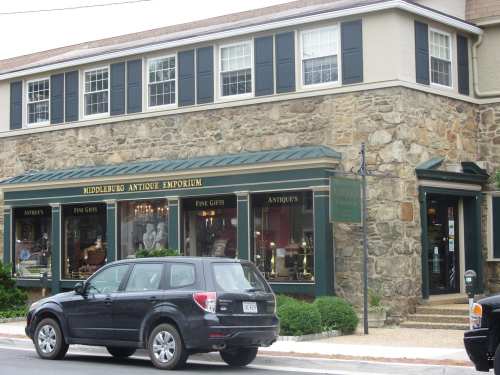 A stone building with a green awning, featuring antique displays in windows and a parked black SUV in front.