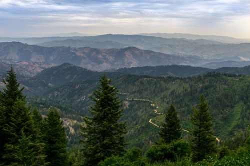 A scenic view of rolling mountains and valleys, with lush green trees in the foreground and a cloudy sky above.
