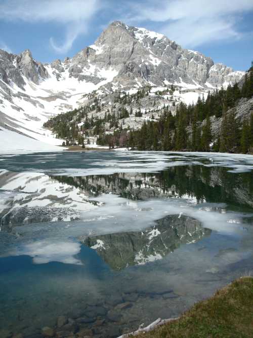 A serene mountain lake reflects snow-capped peaks and lush greenery under a blue sky with scattered clouds.