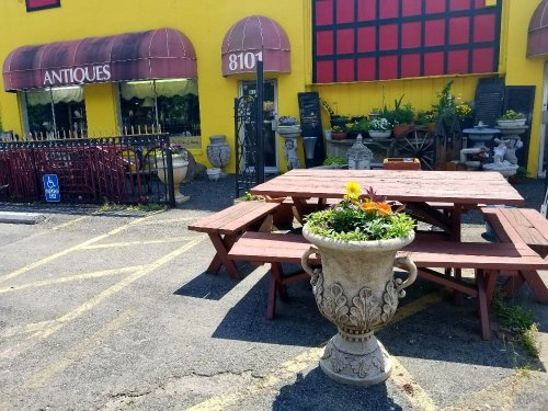 A vibrant yellow antique shop with a red awning, featuring a large flower pot and picnic tables outside.