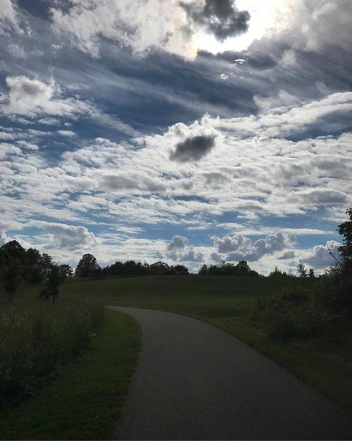 A winding path through a grassy area under a sky filled with clouds and sunlight peeking through.