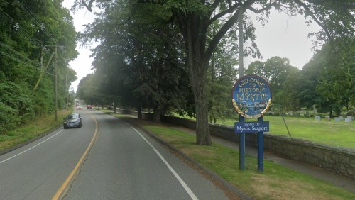 A road sign welcomes visitors to Historic Mystic, with trees lining the street and a cemetery visible in the background.