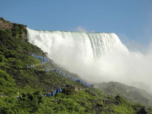 A large waterfall cascades down rocky cliffs, with tourists in blue ponchos observing from the lush green hillside.