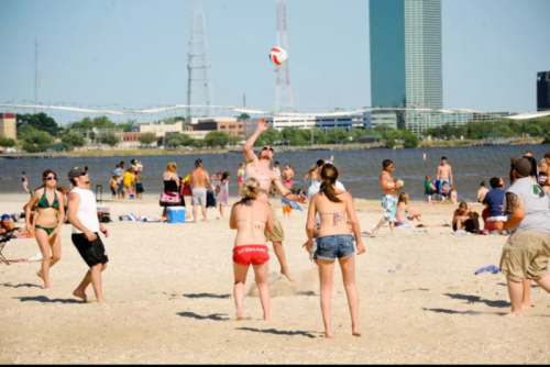 A lively beach scene with people playing volleyball, sunbathing, and enjoying the sunny day by the water.