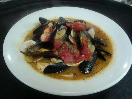 A white bowl filled with a seafood stew featuring mussels, clams, tomatoes, and herbs in a savory broth.