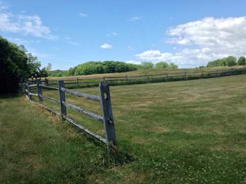 A grassy field with a wooden fence under a blue sky with scattered clouds and green hills in the background.