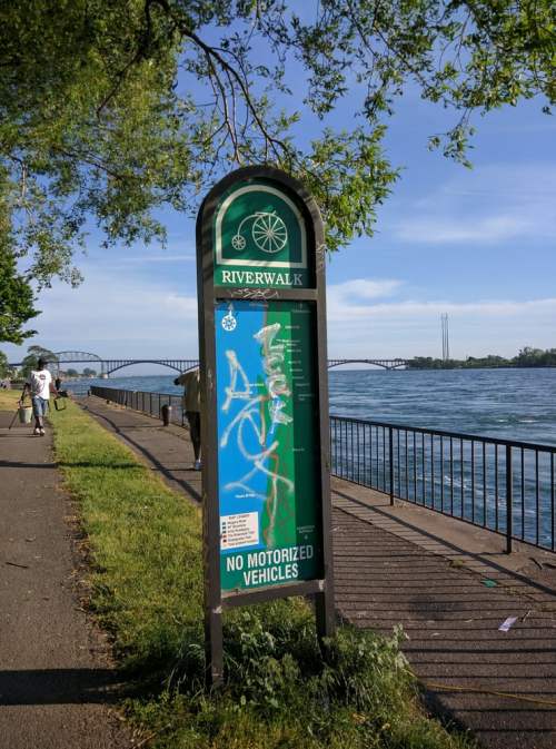 Sign for Riverwalk with graffiti, alongside a river and bridge in the background, surrounded by trees and a walking path.