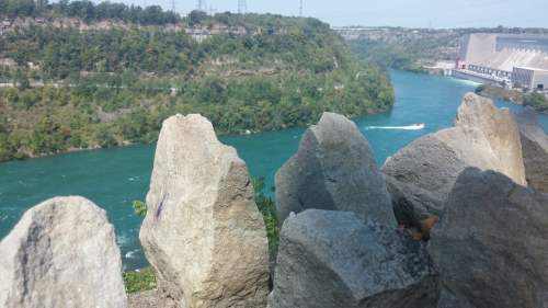 Rugged rocks in the foreground with a vibrant blue river and lush green hills in the background under a clear sky.