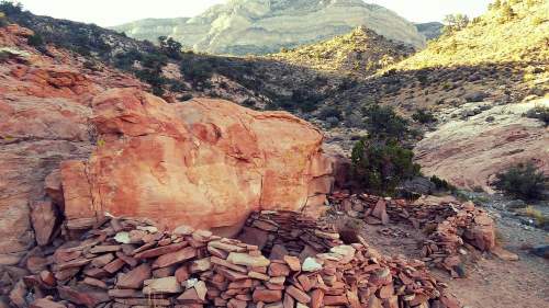 Rocky landscape featuring a stone structure and large boulders, surrounded by mountains and vegetation.