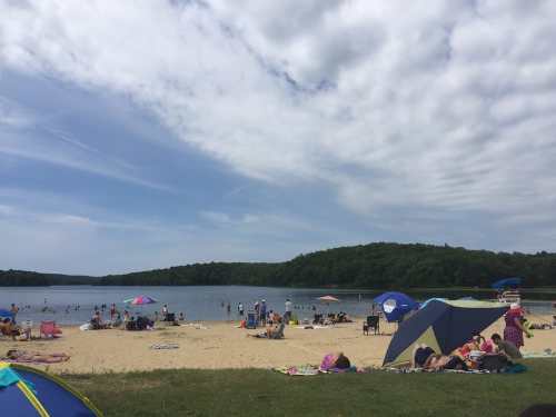 A sandy beach with people relaxing, swimming, and colorful umbrellas under a partly cloudy sky. Green hills in the background.