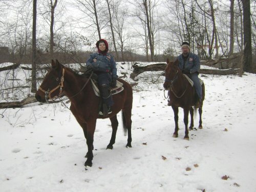 Two people on horseback in a snowy landscape, surrounded by bare trees and fallen branches.