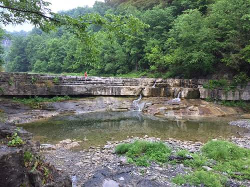 A serene landscape featuring a rocky area with a small waterfall, surrounded by lush green trees and a calm pool of water.