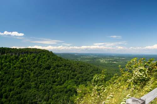 A panoramic view of lush green hills under a clear blue sky with scattered clouds in the distance.