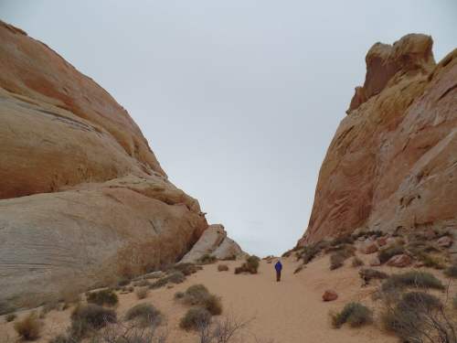 A person walks through a sandy path between towering rock formations under a cloudy sky.